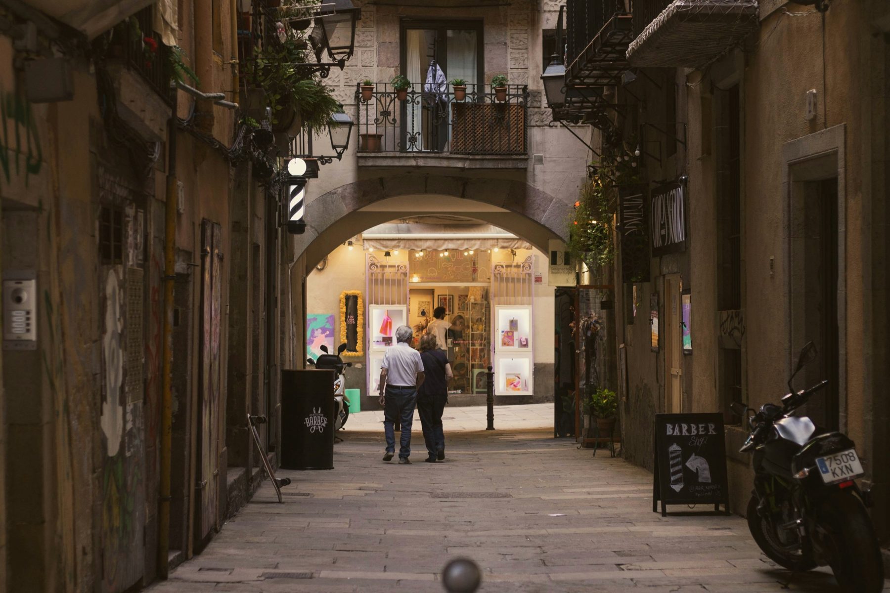 Una parella gran camina per un carreró de Ciutat Vella, a Barcelona, sota un arc i balcons amb plantes.